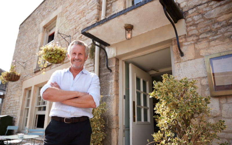 man with gray hair, mustache, and beard wearing white button down with sleeves rolled smiles down toward camera in front of stone residence.