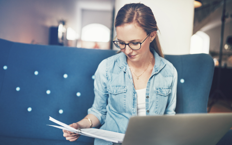 Woman in denim shirt wearing glasses sits on blue couch looking over papers in front of computer. 