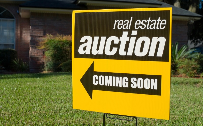 Yellow and black real estate auction coming soon sign in green yard.