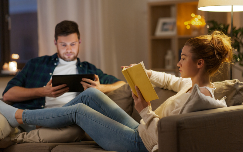 Couple sitting perpendicular on the couch with warm lighting. The man is holding a screen and the woman is reading a yellow book with her leg stretched across his.