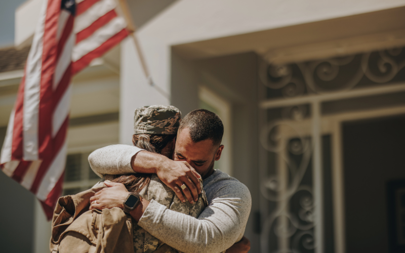 Man wearing long sleeve shirt and watch buries face in female service members in uniform and hugging her in front of home with American flag hanging out front.