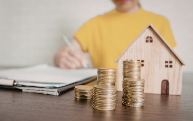 Woman in yellow shirt sits behind desk writing on noted pad with wooden model home and stacks of coins in front of her.
