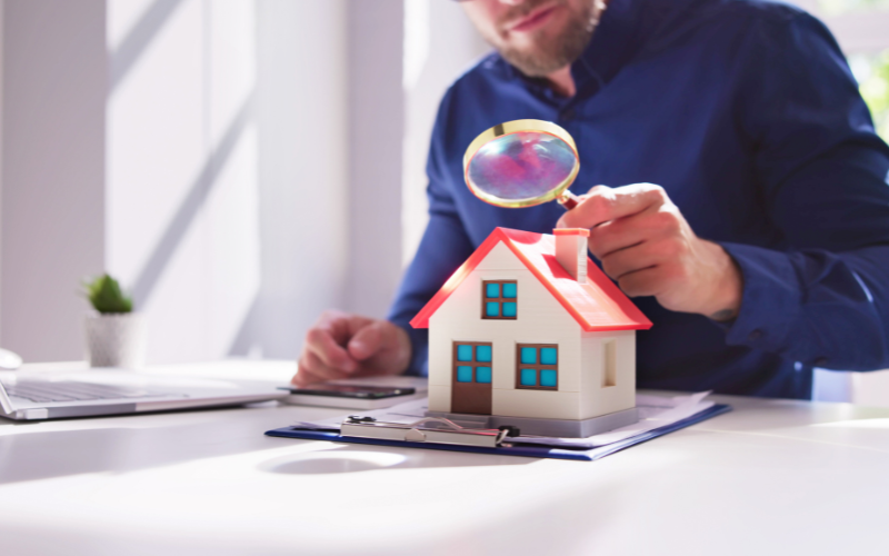 Man in blue shirt holding magnifying glass over model home for home inspection.