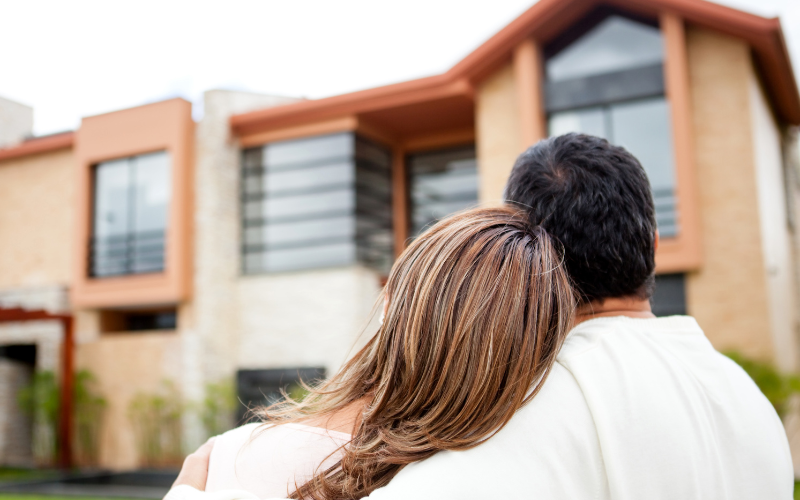 Woman with long hair rests her head on man's shoulder as they stand looking at light brick colored modern home.