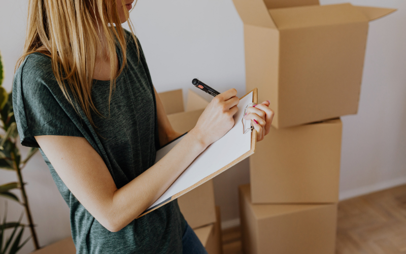 Girl in green shirt stands among moving boxes writing on clipboard with marker. 