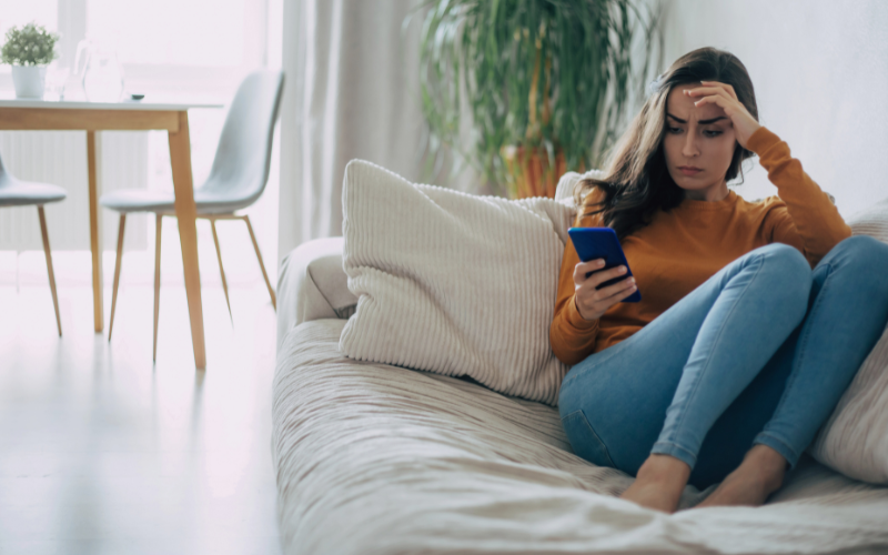 Woman wearing jeans and orange long-sleeve shirt sits in corner of beige couch with hand on her forehead while looking upset at phone in hand.