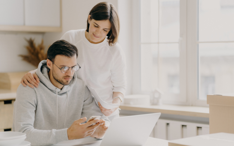 Woman standing over man with phone looking at computer.