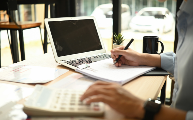 Man wearing black watch types on calculator and writes on paper next to computer sitting at desk by large windows. 