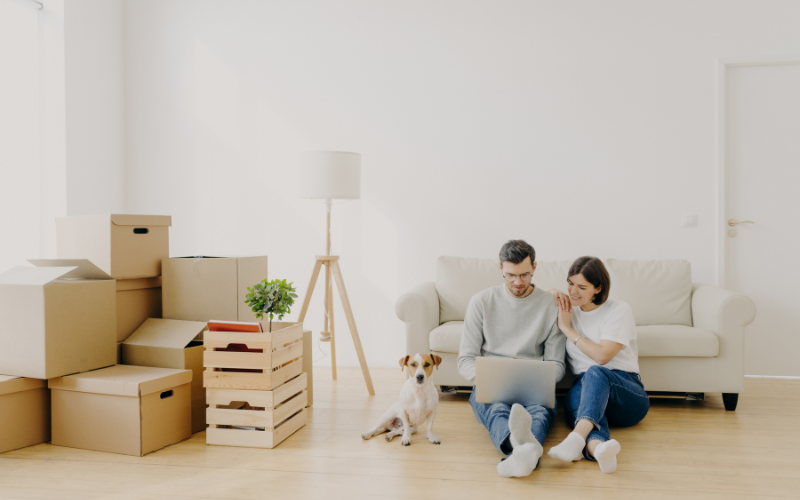 Couple wearing denim jeans sit next to dog on wooden floor in front of the couch while smiling at laptop with moving boxes.