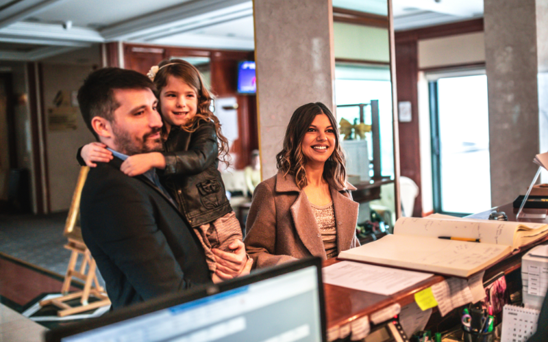man holding child and luggage and woman checking into hotel front desk. 