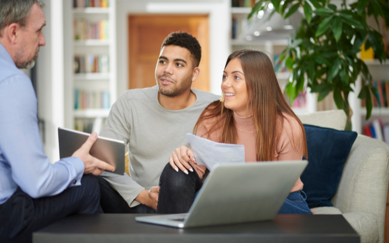 Man and woman sitting across coffee table talking to man with tablet and laptop.