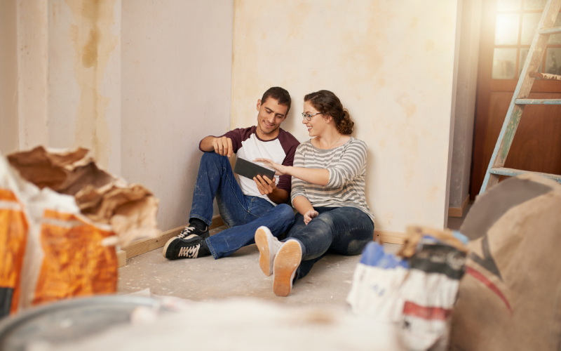 Couple wearing denim sit casually on floor leaning against wall in room under renovation while looking at tablet in hand.