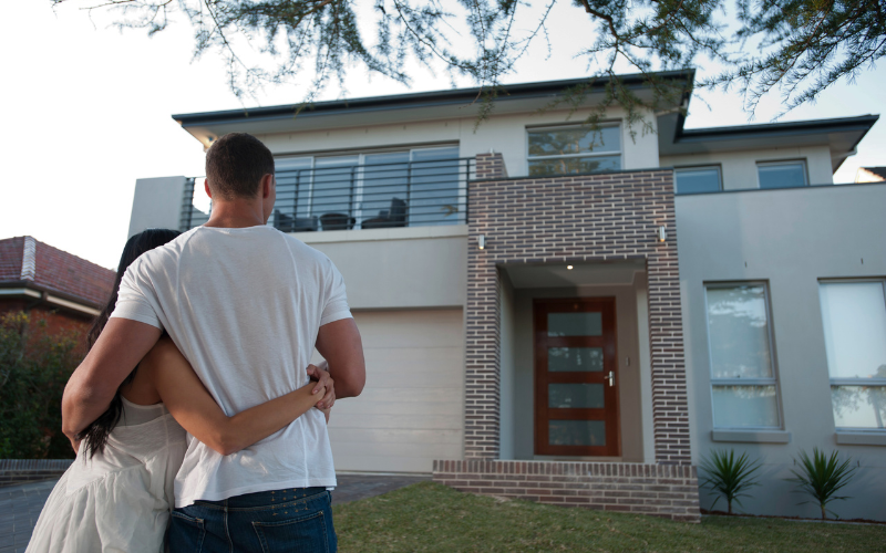 Man in white t-shirt stands with woman's arms wrapped around his waist as they look at modern home with brick details, large windows, and black railings. 