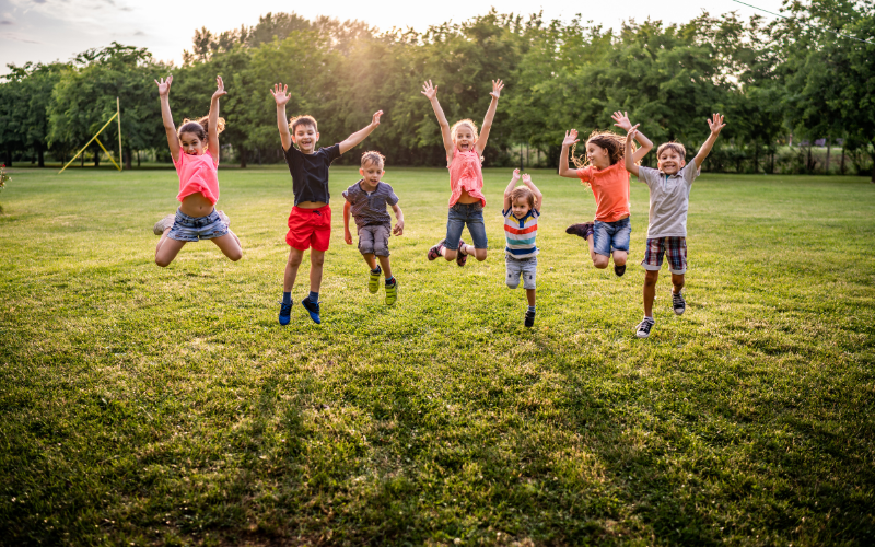 Group of happy kids jumping up together with arms raised in green field.