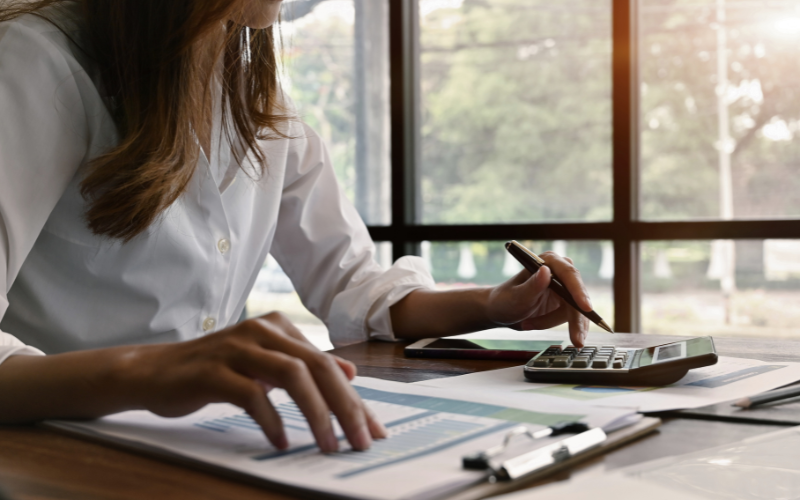Woman wearing a white button-down shirt sits at desk next to window with soft sunlight shining in. She has a pen in her hand, a calculator sitting close, and a clipboard with financial info on it.