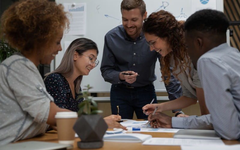 Men and women work professionals in business attire gather around table together smiling and engaging in paperwork sitting in front of them.