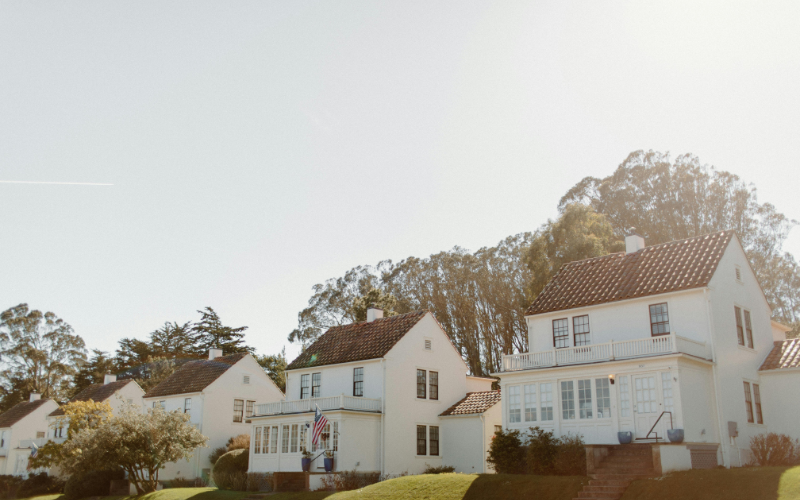 Row of large white houses sitting on small hill with mature trees behind them and an American flag hanging out front. 