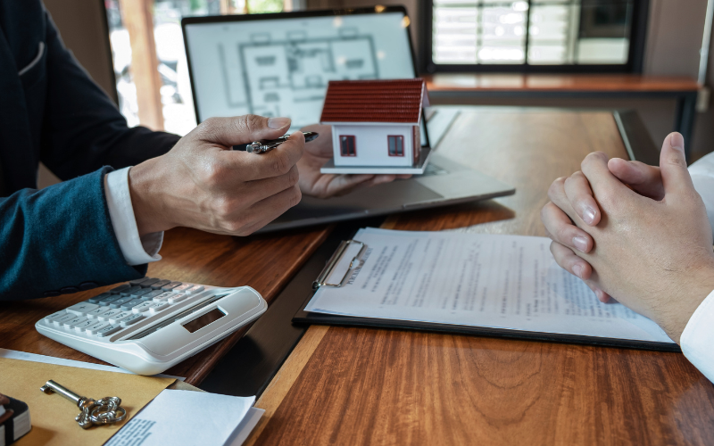 Man wearing blazer with white shirt holds up model house with red roof and points to it with a pen to person sitting across the desk from him. In front of them is a clipboard with paperwork, a computer screen with home layout, a key, and a calculator.