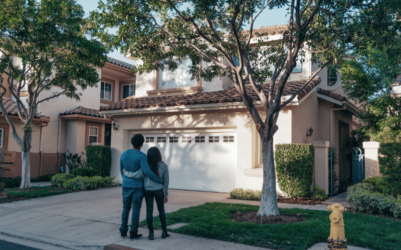 Couple wearing blue stand with their arms around each other looking at stucco two-story home with tile roof and a landscaped green yard.