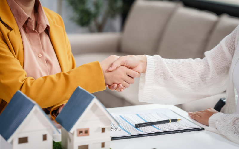 Woman in yellow blazer shaking hands with woman in white across table with model homes and closing agreement on table.