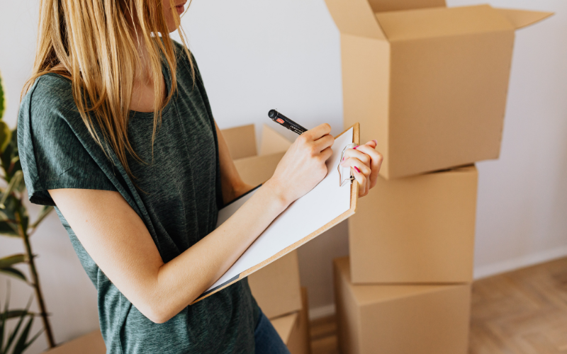 Woman in green shirt writes on clipboard she's holding in front of stacked moving boxes.