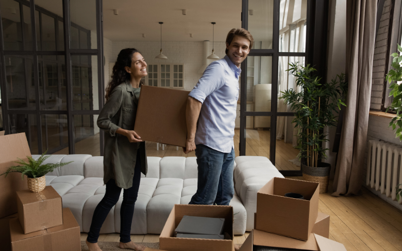 Man in blue button down holds moving box from behind while woman in green button down smiles and helps him carry it into room with other moving boxes on the floor.