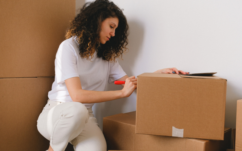 Woman wearing white shirt squats to write on moving boxes with red marker.