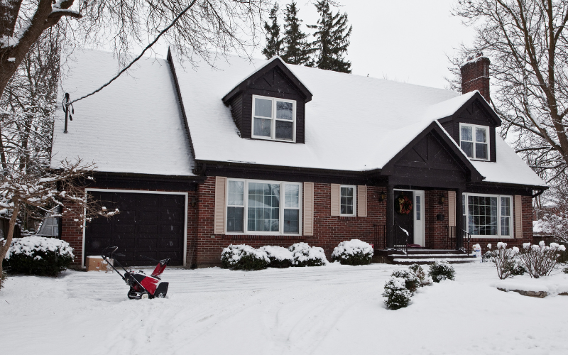 Brick home with dark siding sits covered in snow with red snow blower sitting in driveway.