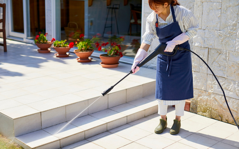 Woman wearing gloves and a blue apron, pressure washes tiled patio outside home. 
