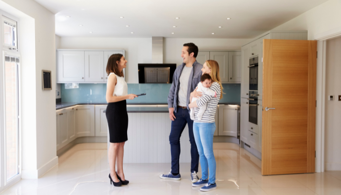 Female property manager/landlord wearing black skirt and heels smiles as she welcomes couple with baby into rental home with shiny kitchen and white floors.