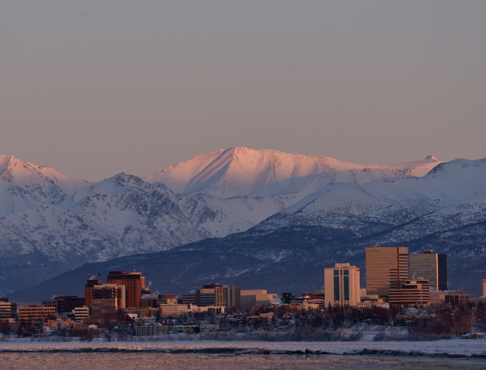 Anchorage, Alaska skyline with a pinkish hue over the blue mountains and downtown buildings. 