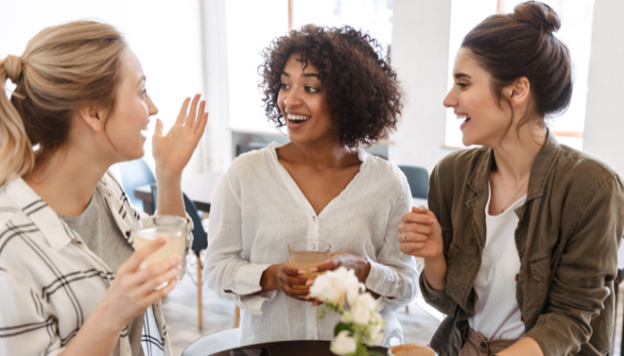 Three women wearing white shirts sit around small table with white roses on it and smile and talk happily to one another.