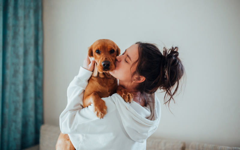 Photo of woman with hair in bun kissing golden puppy in her arms. 
