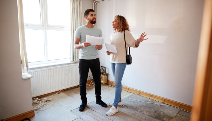 Man and woman standing in bare-bones room holding papers and gesturing around room. 