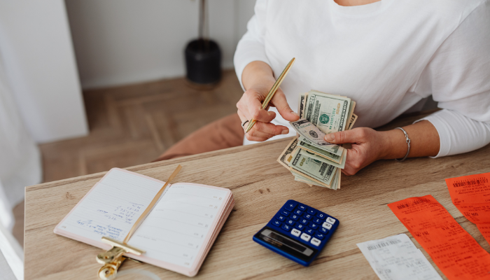View of woman in white shirt thumbing through cash with calculator, notebook, and lists sitting in front of her.