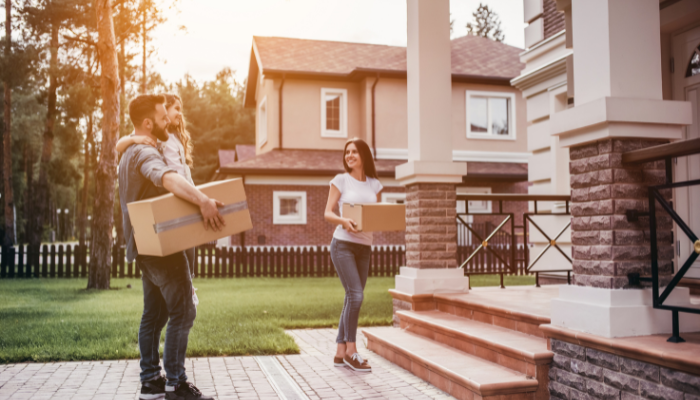Woman holding moving box into house but stops to look back at man with moving box behind her carrying child.