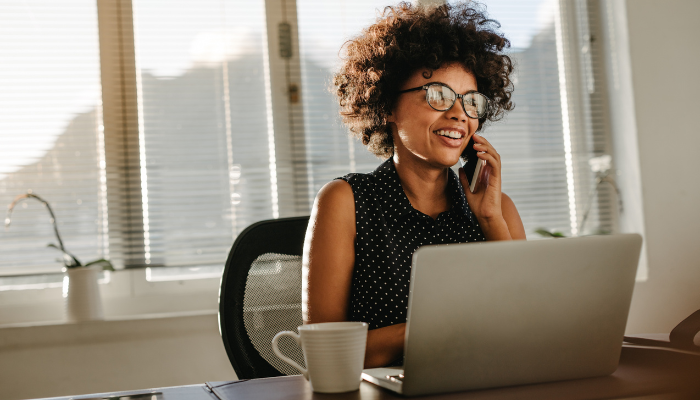 Woman wearing glasses and polka dot sleeveless shirt smiles as she talks on cellphone with computer and coffee mug in front of her and sun shining through windows behind her. 