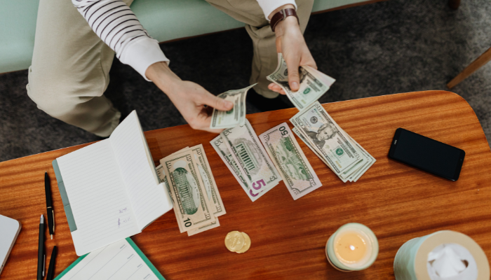 Person in striped long sleeve shirt and khaki pants counts and sorts money on wooden coffee table. 