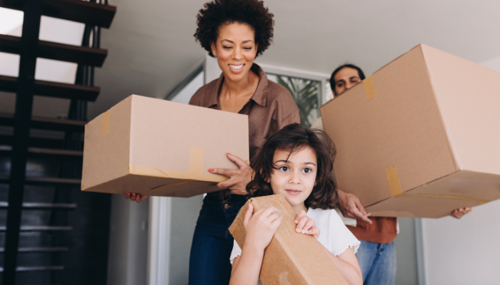 Family of three smiling as they carry moving boxes through house.