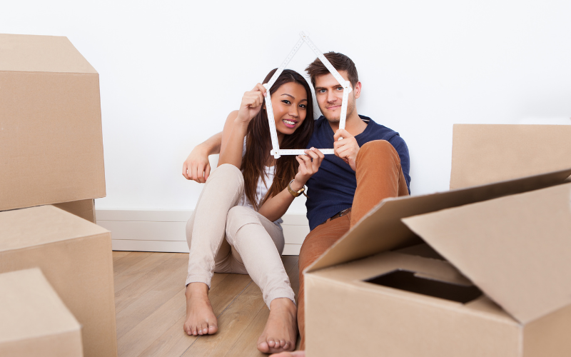 Man and woman sitting together on the floor among moving boxes smiling through white outline of house .