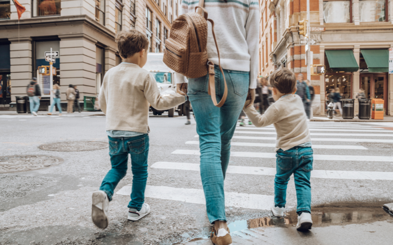 Mom and two boys all wearing beige sweaters and jeans, walk across the crosswalk in city.