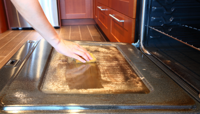 Person scrubbing the interior oven window in kitchen with red-tone wood cabinets. 