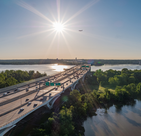 View of traffic on the Wilson Bridge crossing the Potomac River in DC with airplane flying, trees growing in the water alongside the bridge, and sun shining bright.