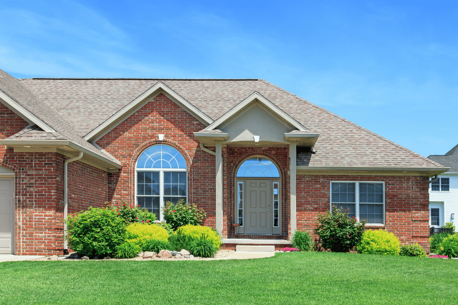 one level red brick home with lush green lawn and bushes on a sunny day with blue sky